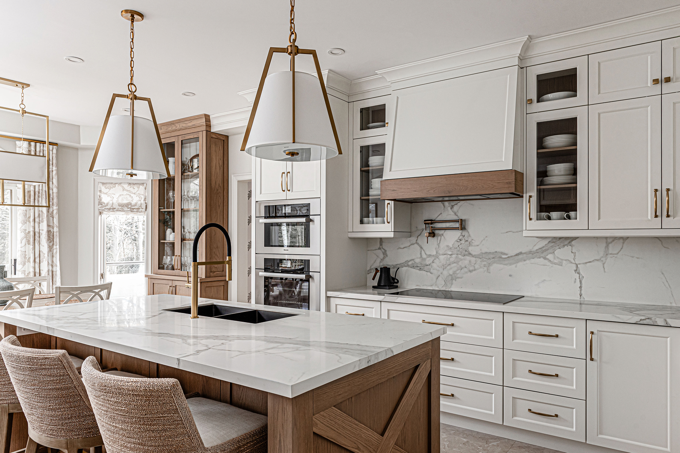 This transitional kitchen blends soft gray cabinetry with warm wood tones. A marble-topped island anchors the space, complemented by brass fixtures, elegant pendant lighting, and a sophisticated marble backsplash throughout.