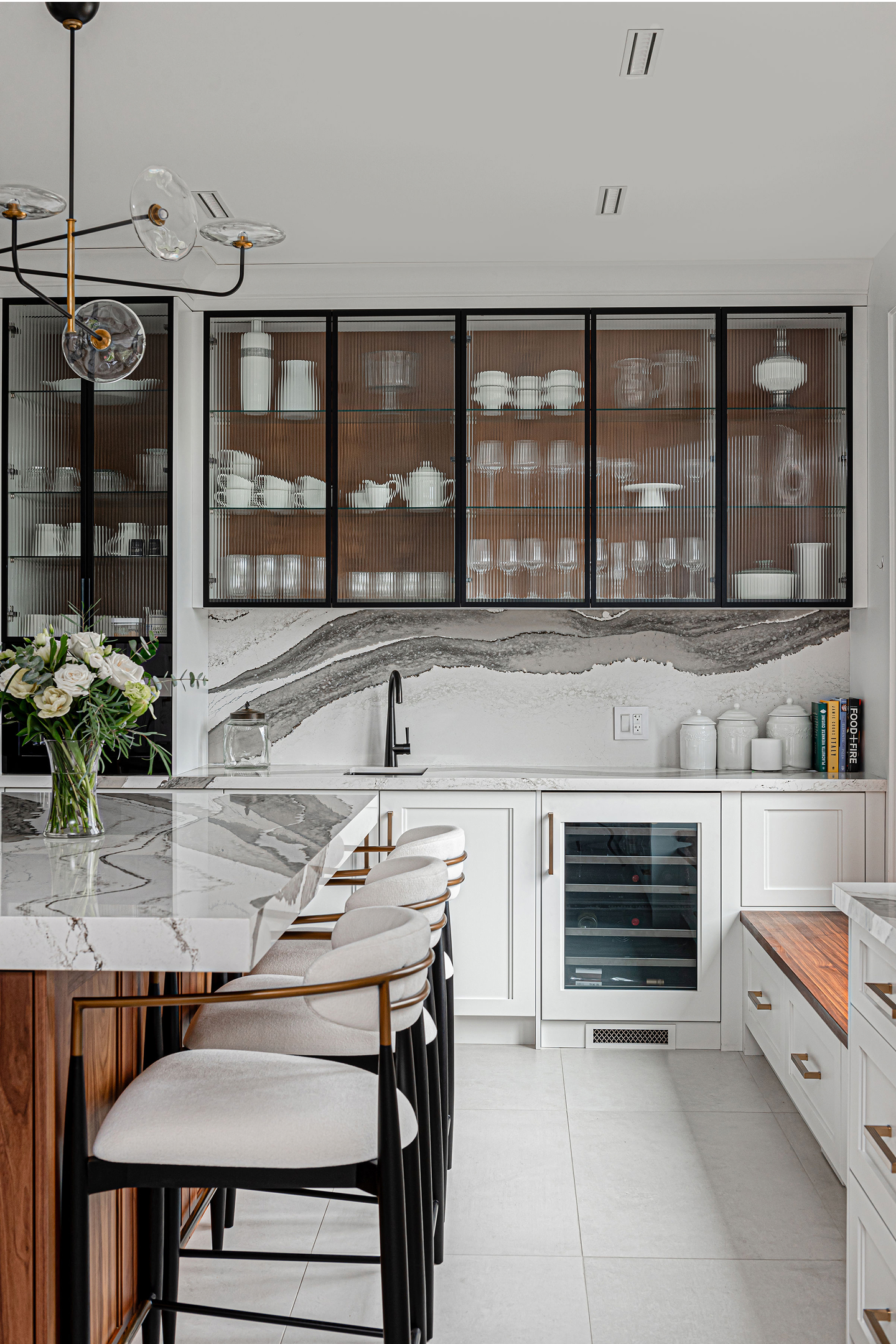 This elegant kitchen showcases stunning gray marble with dramatic veining throughout the island and backsplash. Floor-to-ceiling glass-front cabinets with ribbed backing display white dinnerware, while white cabinetry, integrated wine storage, and a sculptural chandelier complete this sophisticated, gallery-like space.