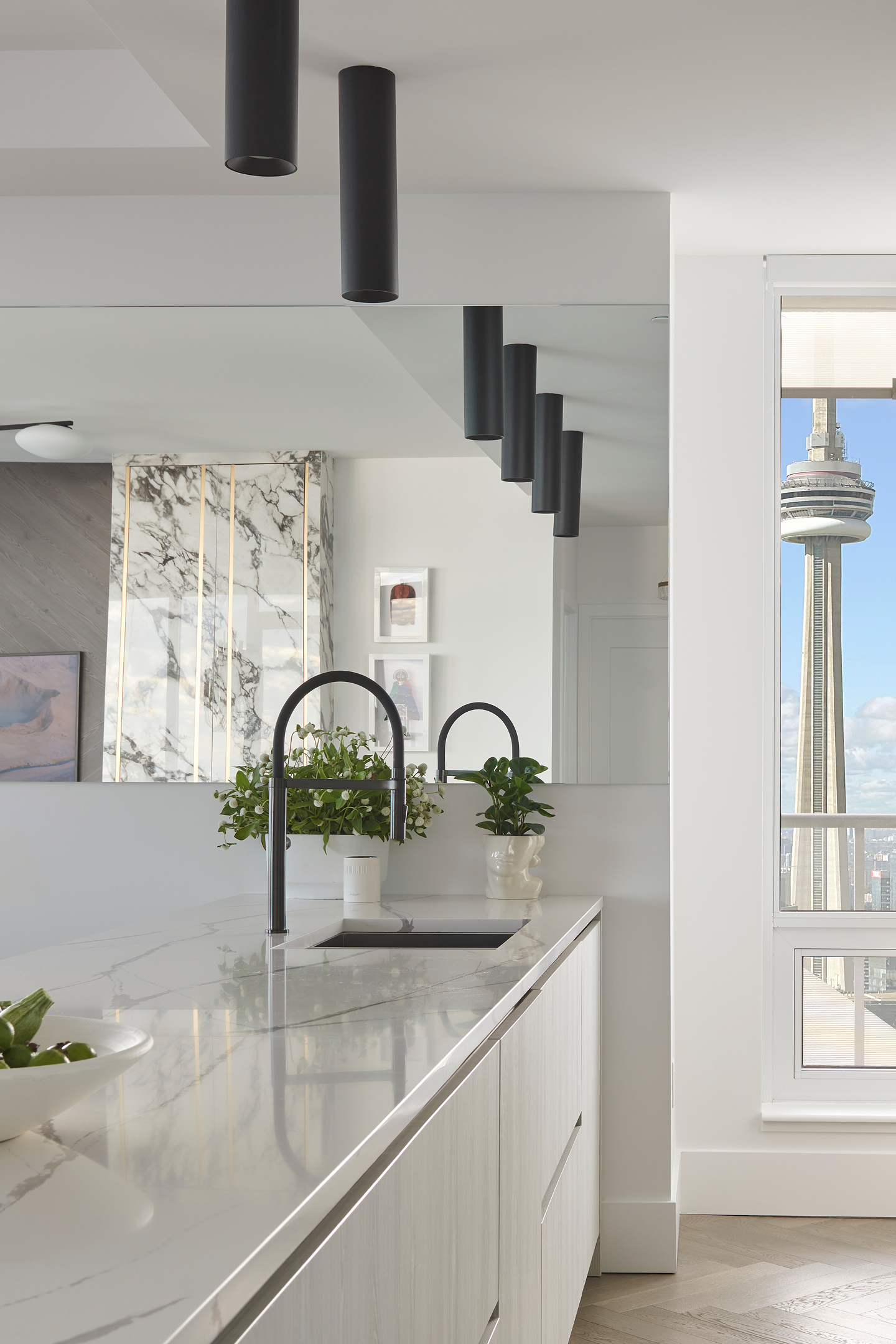 This minimalist modern kitchen features pristine white cabinetry with luxurious marble countertops and a matching statement backsplash. Sculptural black cylindrical pendant lights create dramatic visual interest, while a sleek black faucet and city views add contemporary urban sophistication.