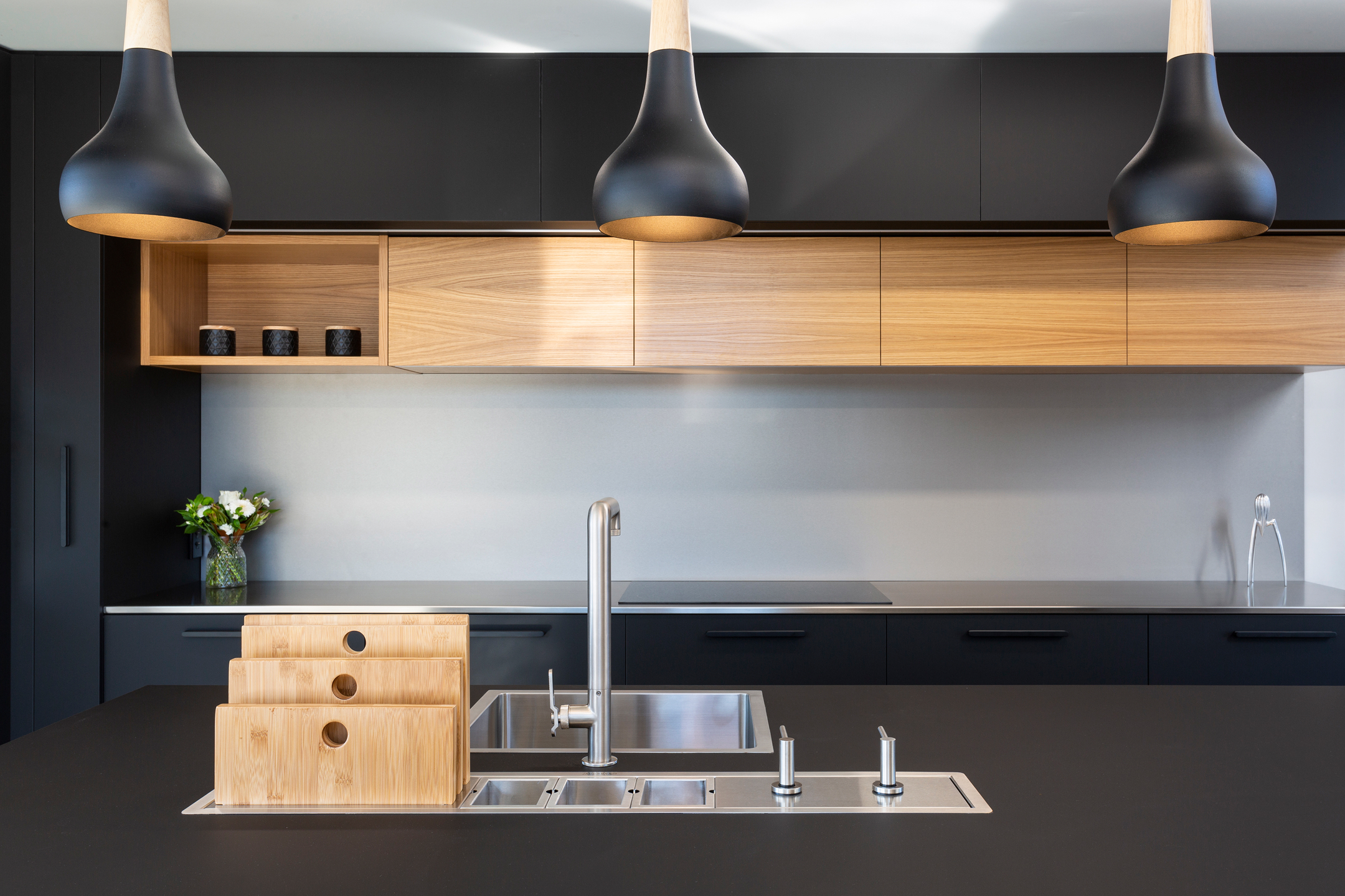This sleek modern kitchen features dramatic black lower cabinets contrasted with warm wood upper storage. Three sculptural pendant lights illuminate the stainless steel countertop and integrated sink, creating a minimalist yet functional contemporary design.