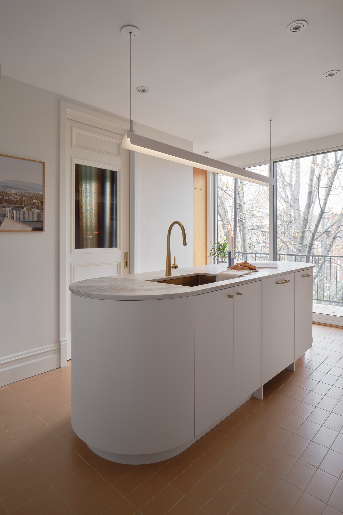 This airy minimalist kitchen features a sculptural curved island with white cabinetry and pale stone countertops. A brushed brass faucet adds warmth, complemented by modern linear pendant lighting. Floor-to-ceiling windows flood the space with natural light, creating a serene, contemporary atmosphere.