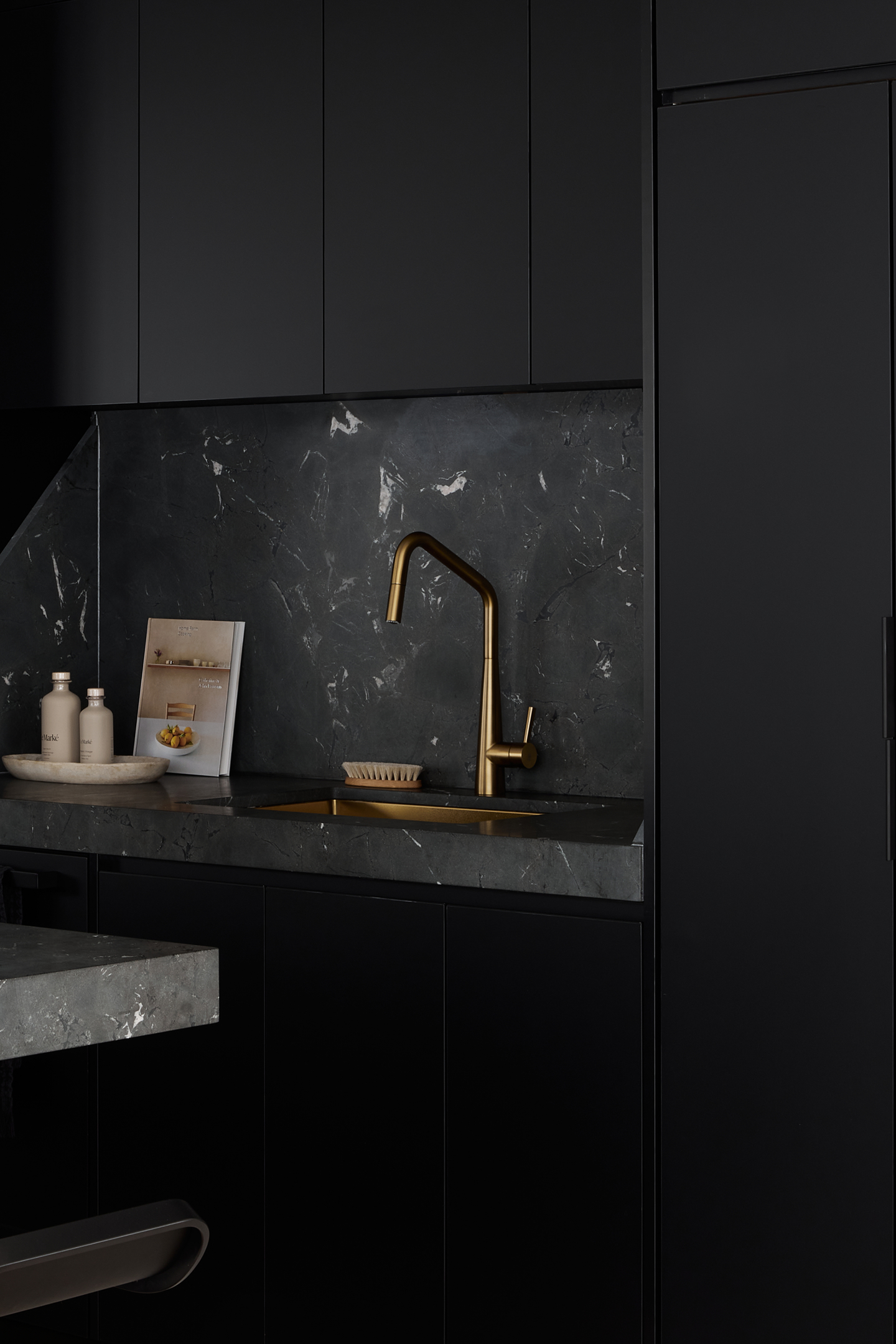 This dramatic all-black kitchen creates bold sophistication with matte cabinetry and dark marble countertops featuring subtle white veining. A striking brushed gold faucet provides the only metallic accent, creating elegant contrast against the moody monochromatic palette and textured stone surfaces.